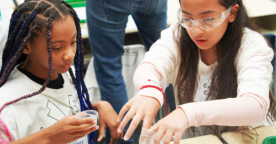 Two elementary school students help each other pour baking powder into a rubber glove for a Science Explorers experiment